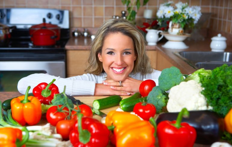 A woman with a perfect smile behind a kitchen counter covered with variety of vegetables.