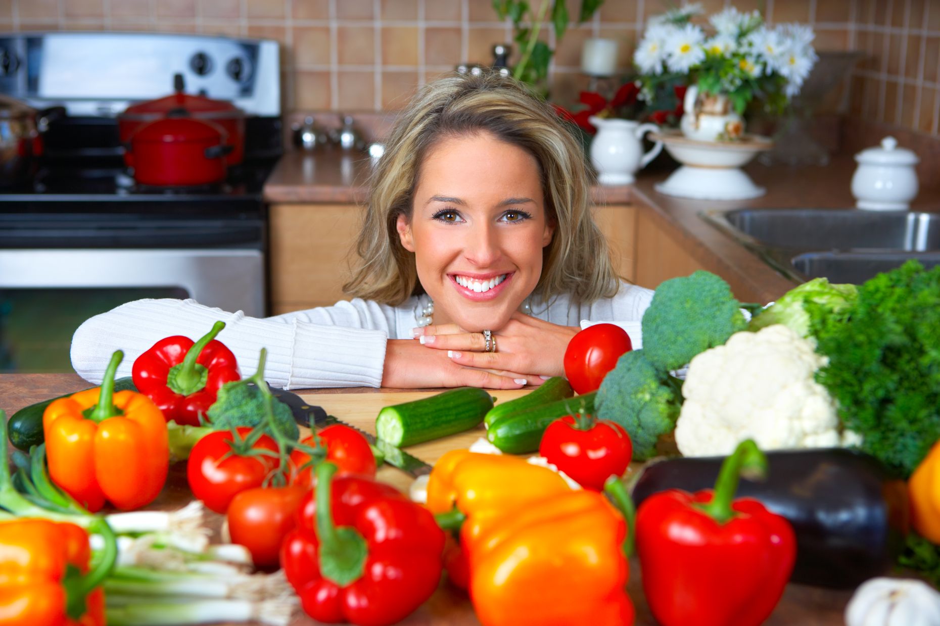 A woman with a perfect smile behind a kitchen counter covered with variety of vegetables.