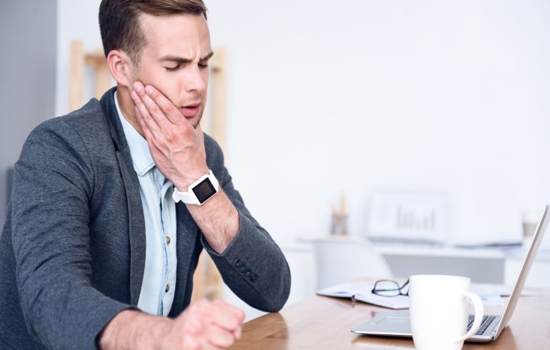 A young businessman sitting at his desk with a laptop and holding his jaw due to pain caused by TMJ syndrome