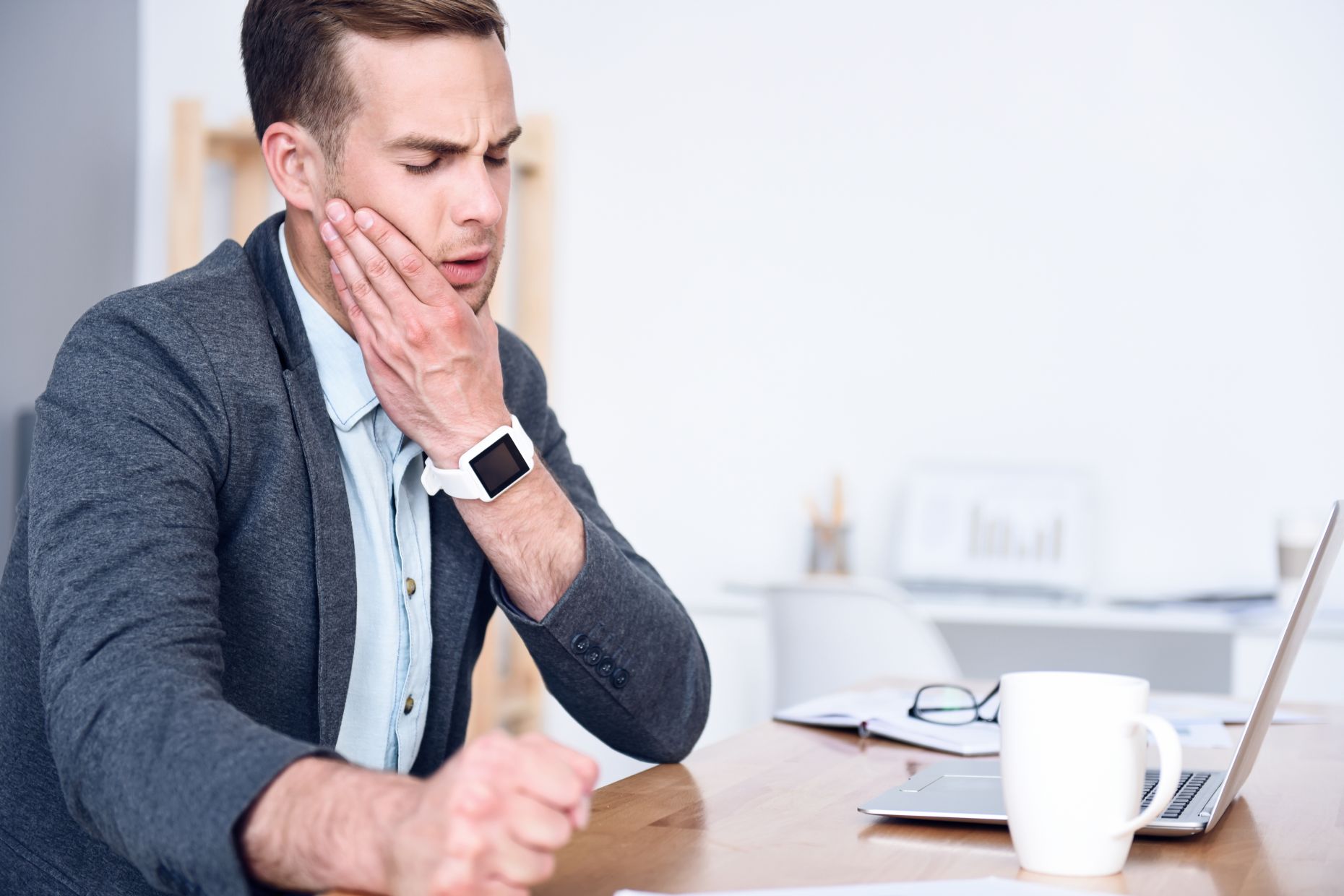 A young businessman sitting at his desk with a laptop and holding his jaw due to pain caused by TMJ syndrome